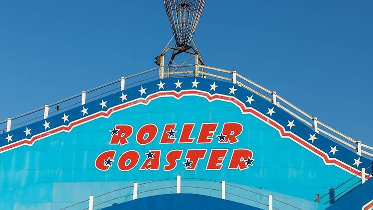 The classic signage of a vintage wooden roller coaster with blue and red American star design against a clear sky — representing historic amusement park heritage